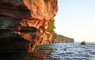 Red sandstone cliffs jutting out over the lake are lit up by the sunset as kayakers paddle away in the distance.