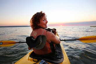 Woman turns around in yellow sea kayak, smiles for camera; sun setting in background.